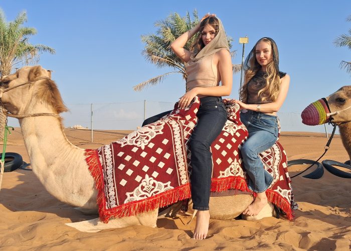Two guests sit on a kneeling camel with a red patterned saddle in Dubai’s desert, blue sky and palm trees behind.
