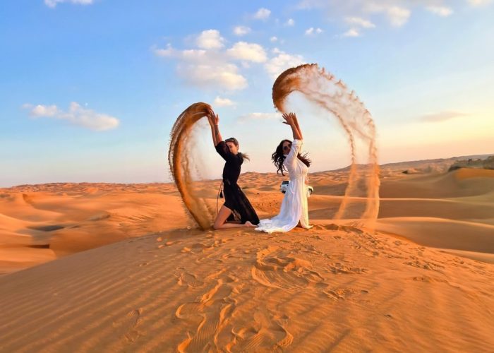 Two guests kneel on a dune at sunset, tossing arcs of sand into the air; vast orange Red Dunes and a white 4×4 in the background.