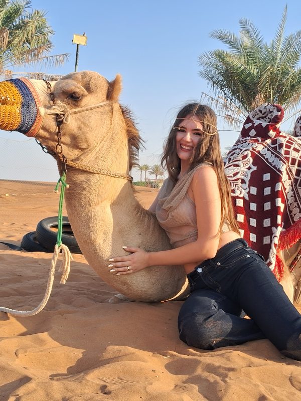 Two guests sit on a kneeling camel with a red patterned saddle in Dubai’s desert, blue sky and palm trees behind.